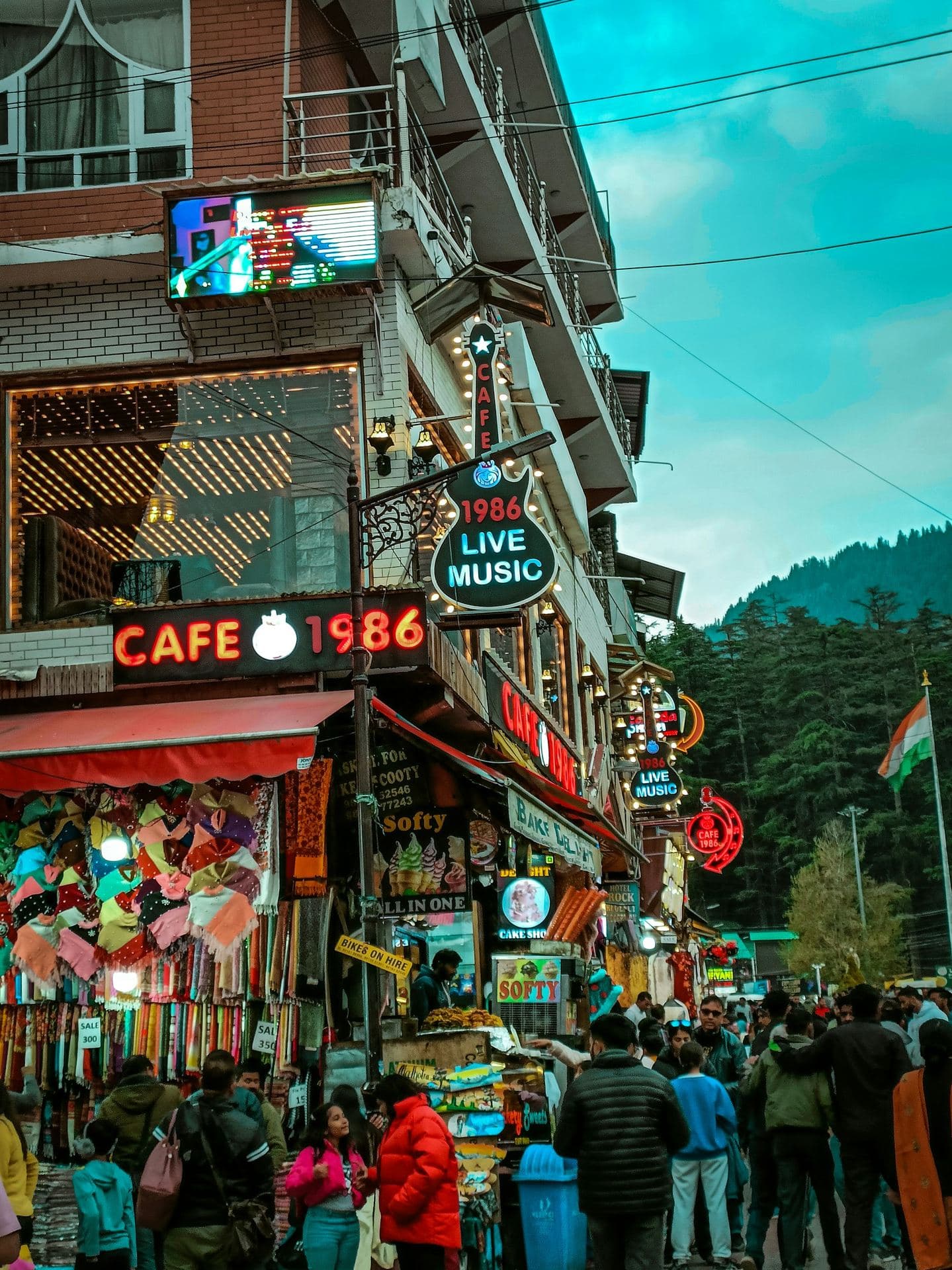 The cafe lane of Old Manali village climbing toward Manu Temple, stone-and-wood houses and apple orchards on the hillside, snow peaks behind