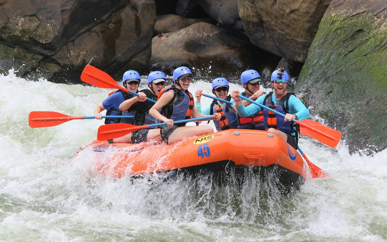 Rafters in a blue inflatable raft riding a white rapid on the snow-fed Beas river near Kullu, forested valley slopes rising behind